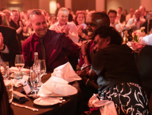 A woman and two men seated at a dinner table at a Gala Dinner. The woman is embracing the younger man, and there is a crowd sitting around them applauding. They are three representatives from the Learning on Country Program.