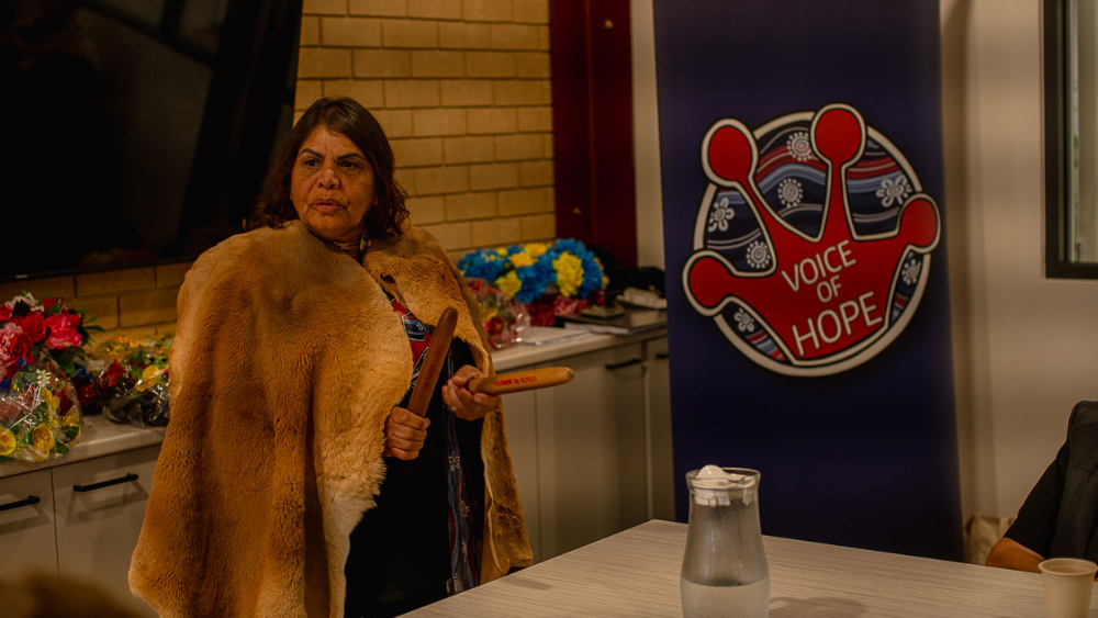 A middle-aged Aboriginal woman stands in front of a banner with a logo reading 'Voice of Hope'. The woman is wearing a cloak made of animal fur and is holding two clapsticks. She looks to be in mid-speech.