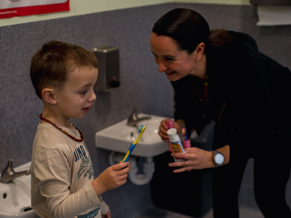 A woman holding a toothbrush and toothpase smiles a child holding his own toothbrush.