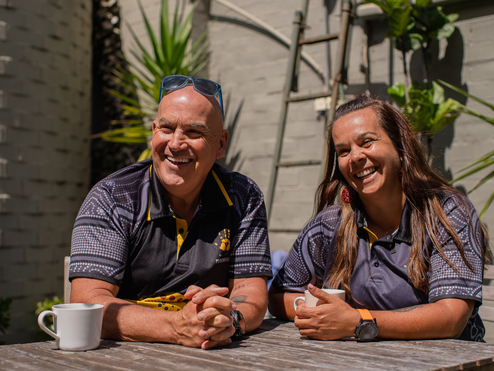 Two people sit at a table outside, in the sun, holding coffee mugs, smiling at something off camera.
